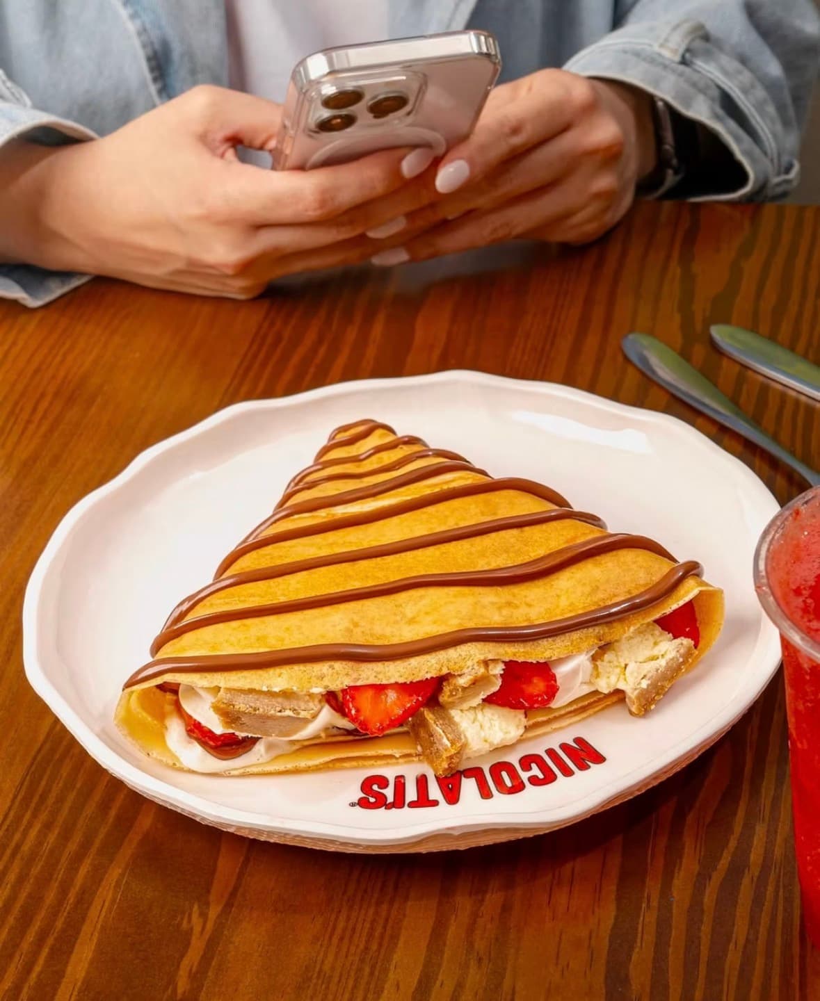 A Nicolati's strawberry-and-cream crêpe drizzled with chocolate on a branded plate, as a customer films it on their phone to post on TikTok.